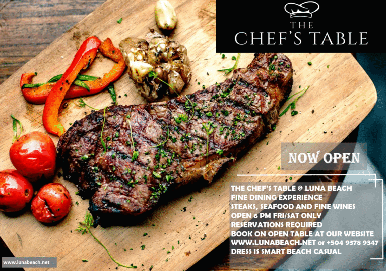 A wooden serving board displays a grilled steak garnished with chopped herbs, accompanied by roasted cherry tomatoes, sliced red bell peppers, green beans, and a roasted garlic bulb, with a black and white promotional overlay for The Chef's Table at Luna Beach in Roatan, Honduras.