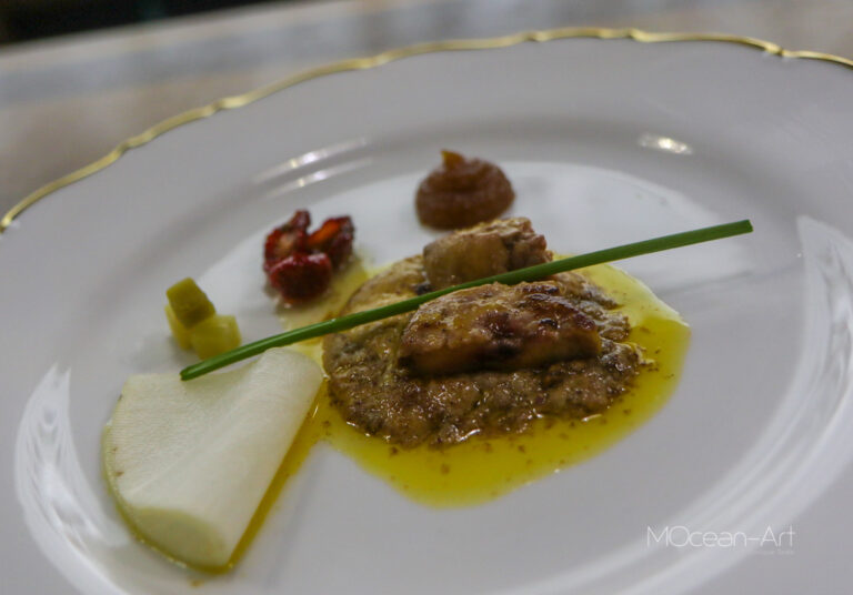 A gourmet dish featuring a piece of meat in a yellow sauce, garnished with a chive, served alongside a slice of pear, small diced vegetables, and dollops of accompaniments on a white plate with gold trim, set against a blurred background.