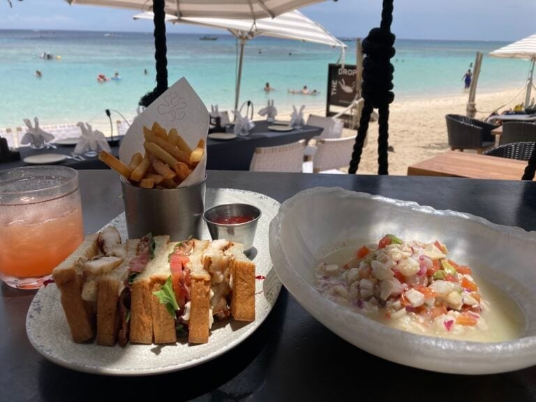 A table set for dining with sandwiches, french fries, and creamy potato salad overlooking the beach and turquoise waters of Roatan, Honduras, with umbrellas and people swimming in the background.