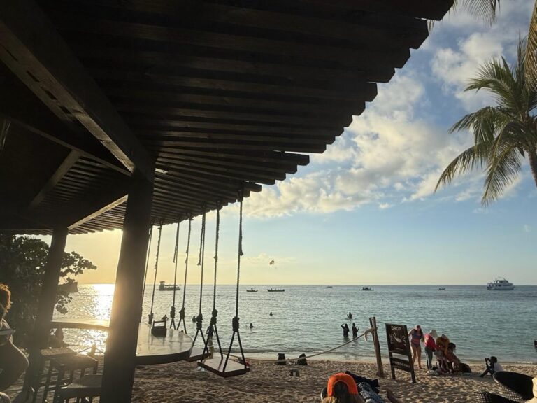 A beachfront scene in Roatan, Honduras, features a shaded outdoor area with swings, a sandy beach with people swimming and relaxing, and the ocean with boats and a boat cruise visible under a partly cloudy sky.
