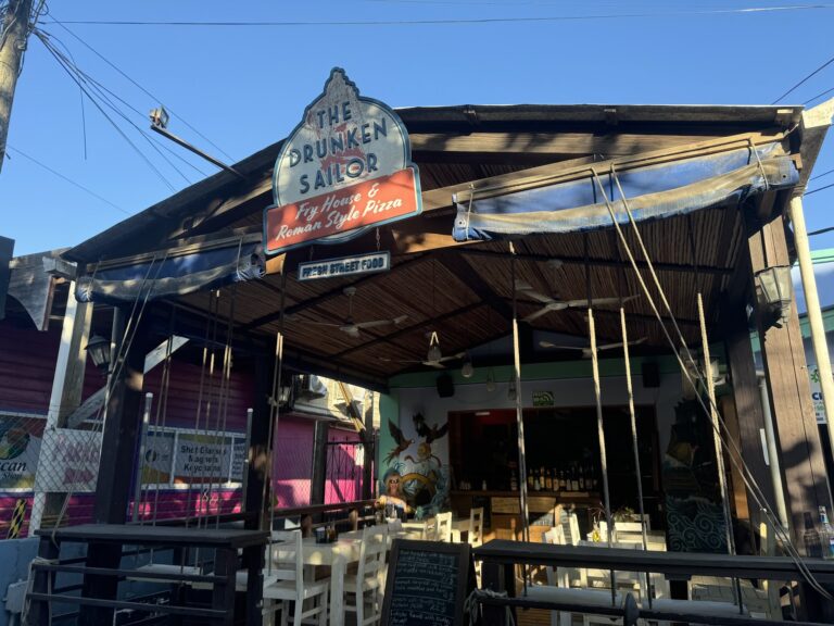A rustic open-air restaurant in Roatan, Honduras, features a sign for "The Drunken Sailor" offering fry house and Roman-style pizza with outdoor seating, wooden decor, and a woman sitting near the tables under a clear blue sky.