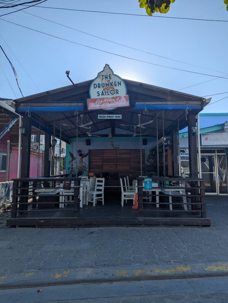 The open-air patio of The Drunken Sailor restaurant in Roatan, Honduras, features white chairs, a wooden deck, and a sign advertising Roman-style pizza and street food, with colorful murals and tropical trees visible in the background.