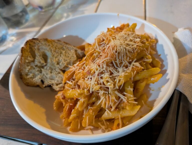 A white bowl of pasta with shredded cheese and a slice of bread is placed on a wooden table, with water glasses and a napkin nearby, in Roatan, Honduras.