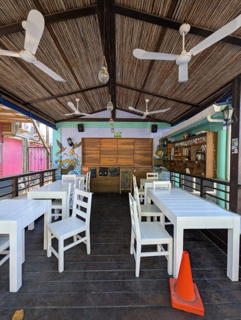 A covered outdoor dining area in Roatan, Honduras, features white tables and chairs, decorative murals on the walls, ceiling fans, hanging light bulbs, and an orange traffic cone on the dark wooden floor.
