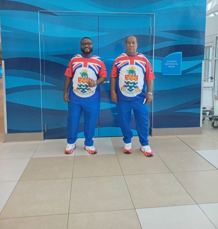 Two men wearing matching Roatan and Honduras-themed jerseys stand in front of a blue wall with wave-like patterns at an airport in Roatan, Honduras.