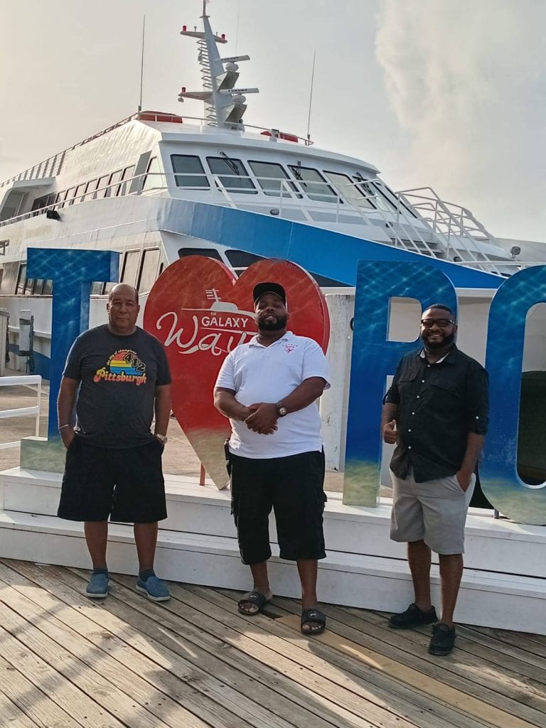 Three men stand in front of a large boat with the logo "Galaxy Wave" on Roatan, Honduras, during daytime, with a prominent "Roatan" sign behind them and the boat's upper deck visible in the background.