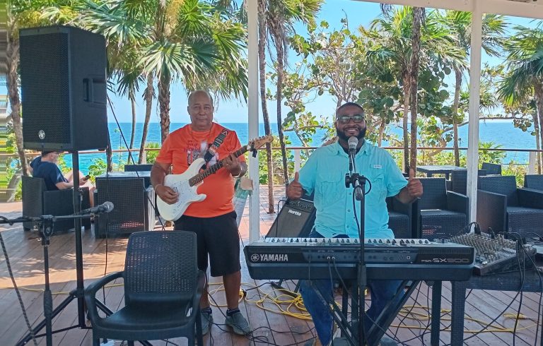 Musicians perform live music outdoors on a balcony overlooking the ocean in Roatan, Honduras, with palm trees and a clear blue sky in the background.