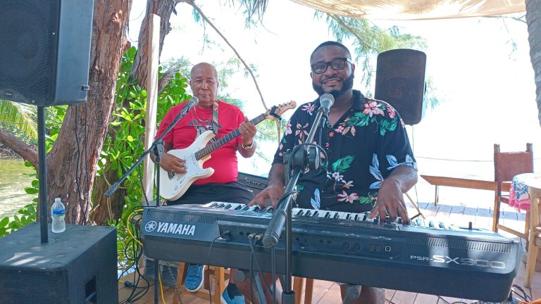 Musicians perform live music on a wooden deck surrounded by trees and tropical greenery in Roatan, Honduras, with one playing a keyboard and the other holding a guitar during an outdoor event.
