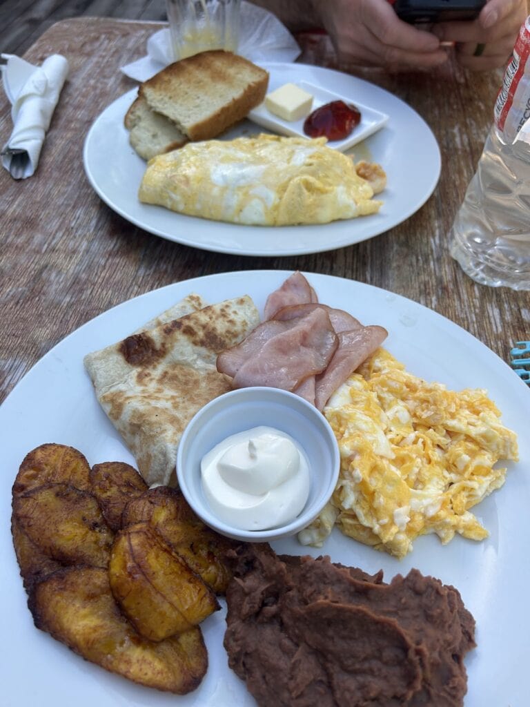 A plate of traditional Honduran breakfast food including scrambled eggs, fried plantains, crispy bacon, and a folded tortilla, served with a small cup of sour cream, on a wooden table in Roatan, Honduras.