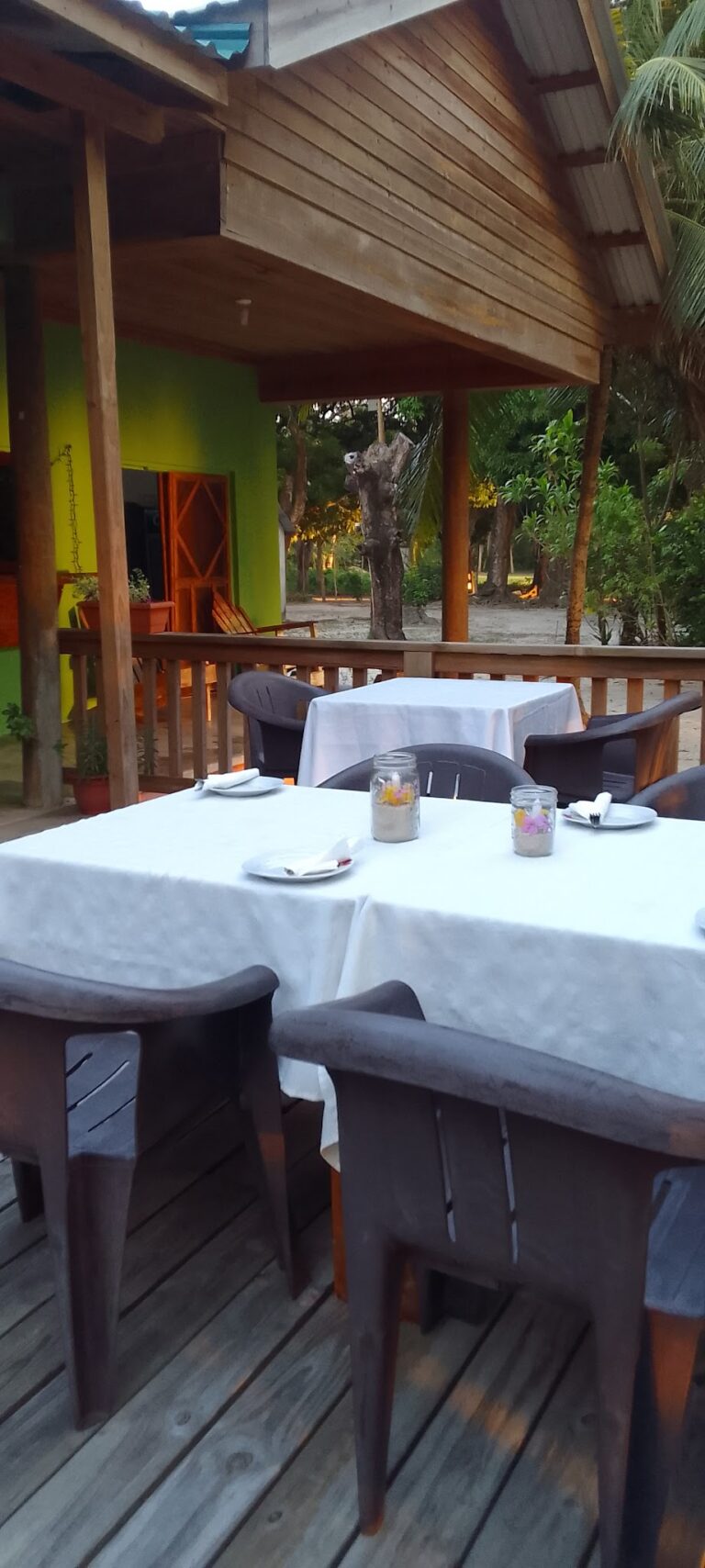 Outdoor dining area with white-tableclothed tables, surrounded by plastic and wooden chairs, set on a wooden deck with a view of trees and lush greenery in Roatan, Honduras.