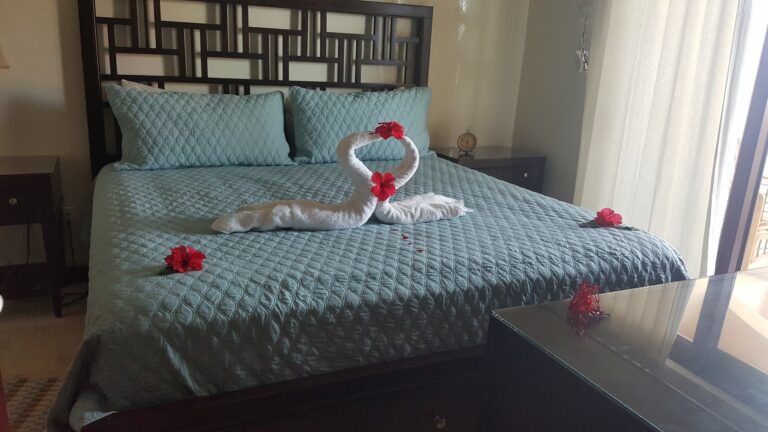 A bedroom in Roatan, Honduras features a bed with a blue quilted bedspread and matching pillows, decorated with a towel art swan and red hibiscus flowers, with natural light coming through a sliding glass door covered by sheer curtains.