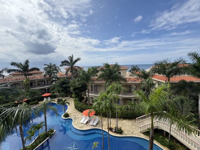 A view of a swimming pool surrounded by palm trees and buildings with red-tiled roofs overlooking the ocean on Roatan, Honduras.