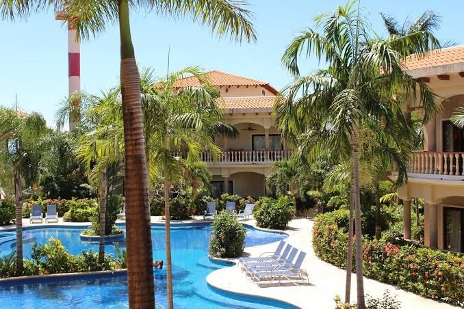 A luxurious resort in Roatan, Honduras features a large freeform swimming pool surrounded by tropical palm trees, lounge chairs, and landscaped gardens, with a multi-story building and balconies overlooking the pool area.