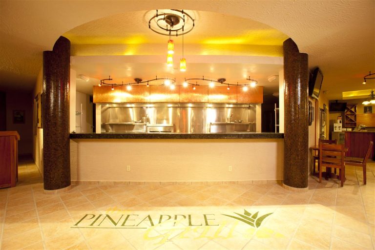 The image shows a warm, inviting restaurant or bar counter in Roatan, Honduras, with a curved wooden and tile design, exposed kitchen area behind the counter, and ambient yellow lighting, featuring the "Pineapple" logo on the tiled floor.
