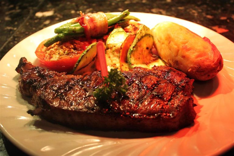 A plated grilled steak served with roasted vegetables, including squash, tomato, green beans wrapped in bacon, and a baked potato, displayed on a white dish for a restaurant in Roatan, Honduras.