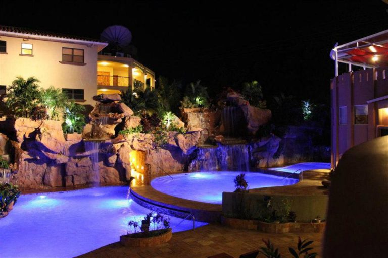 A illuminated swimming pool with waterfalls and rock formations at night outside a hotel or resort in Roatan, Honduras, surrounded by lush tropical plants and a building with balconies and modern architecture.