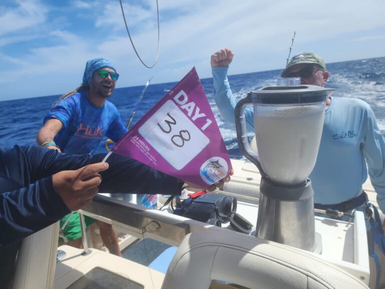 Two men are celebrating on a boat in the clear blue waters near Roatan, Honduras, with one holding a pink race flag and the other raising his fist, while a blender with a white beverage is on the boat's console.