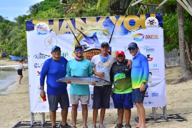 Five men in colorful fishing attire pose on a beach in Roatan, Honduras, holding a large fish with a backdrop featuring logos and banners from a fishing tournament and the scenic shoreline with palm trees and boats.