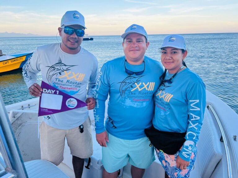 Three people wearing matching blue "Roatan International Fishing Tournament" shirts and white hats stand on a boat against an ocean backdrop in Roatan, Honduras, with a distant shoreline and calm waters visible.