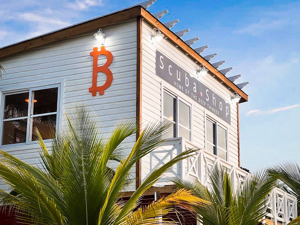 The two-story Cubashop building in Roatan, Honduras, features white wooden siding, large windows, a prominent orange Bitcoin symbol, and a sign that reads 'HOME OF THE DEEP THINKER,' with tropical palm fronds in the foreground and a clear blue sky overhead.