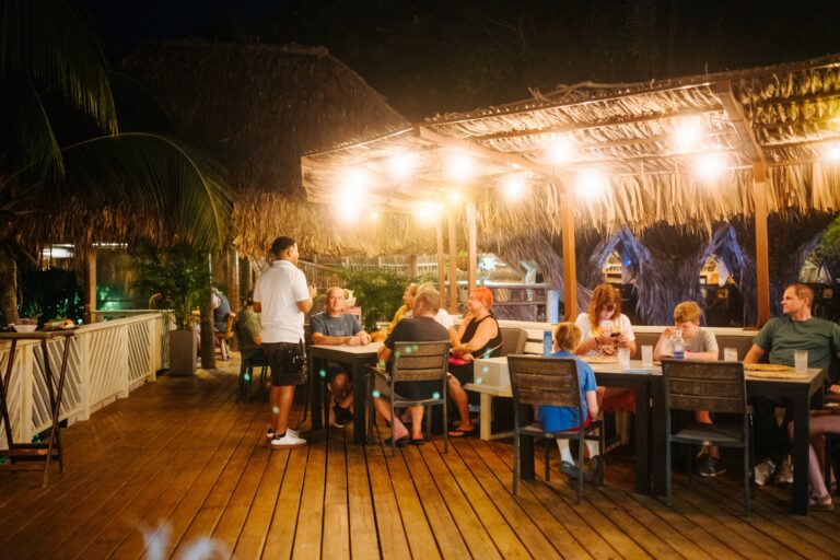 People dining and socializing under a thatched-roof outdoor restaurant on Roatan in Honduras at night, illuminated by string lights and surrounded by tropical foliage.