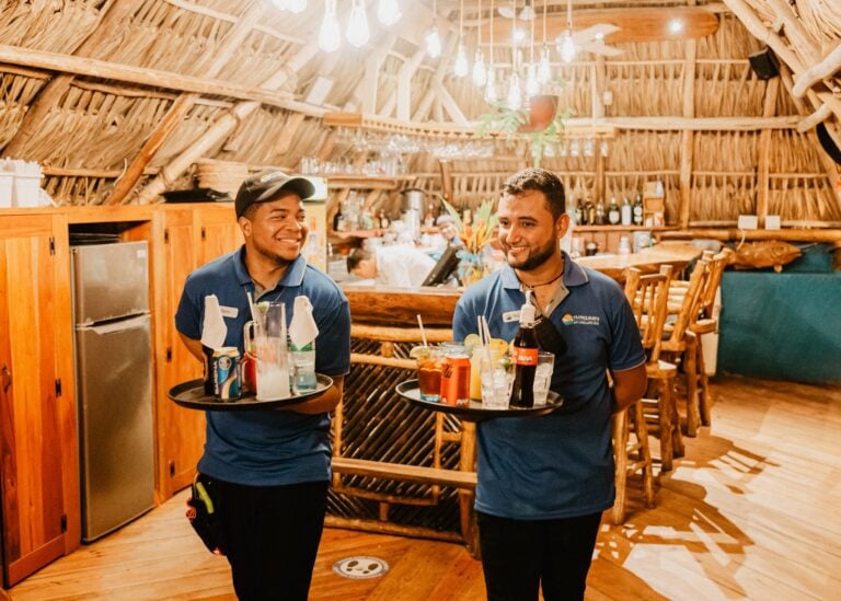 Two smiling waitstaff members are serving drinks inside a rustic, open-concept restaurant with woven bamboo walls and a wooden ceiling in Roatan, Honduras.
