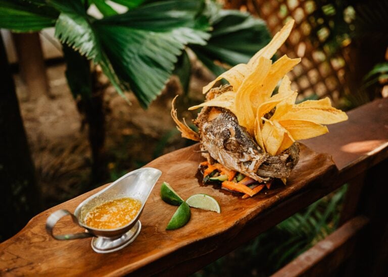 A whole fried fish served on a wooden platter with plantain chips, lime wedges, and a side of yellow sauce at a restaurant in Roatan, Honduras.