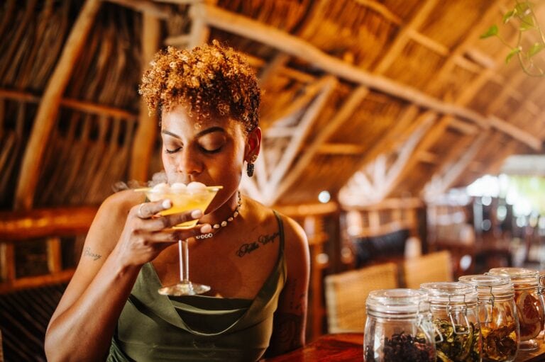 A woman with curly auburn hair and tattoos on her arms enjoys a drink in a rustic, thatched-roof setting at a business in Roatan, Honduras, with glass jars filled with herbs and spices on a wooden counter nearby.