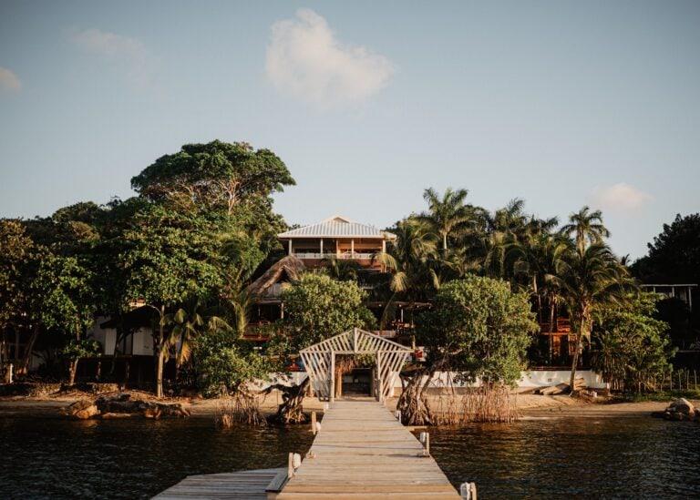 A wooden pier extends into the water leading to a beachside resort with lush tropical trees and buildings on Roatan, Honduras.
