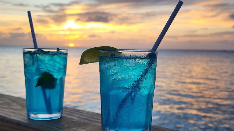 Two blue tropical cocktails with lime wedges and straws are shown on a wooden surface against a sunset over the ocean in Roatan, Honduras.