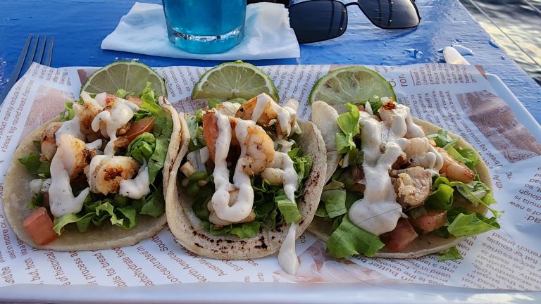 Three shrimp tacos topped with lettuce, white sauce, and lime wedges are served on a paper-lined tray at a restaurant in Roatan, Honduras.
