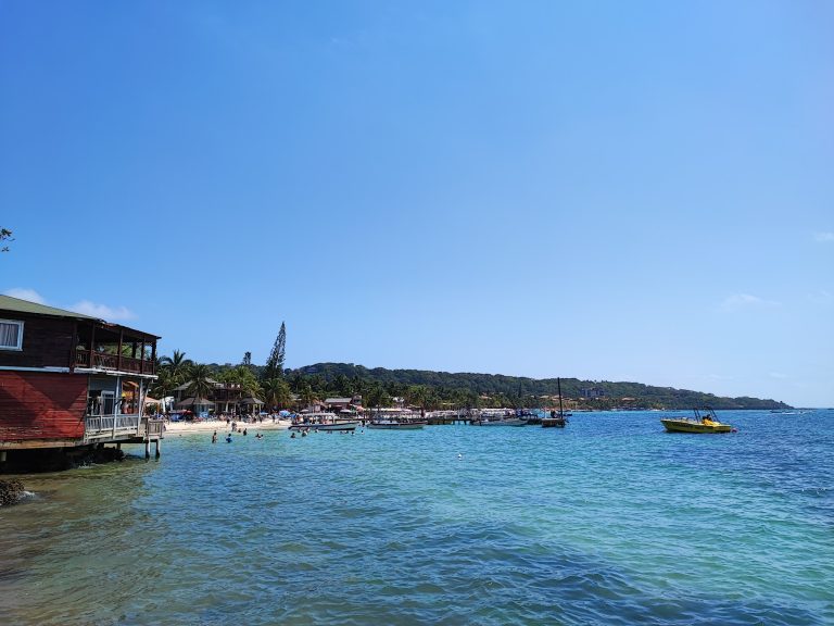 A view of the coastline in Roatan, Honduras, showing overwater bungalows, boats docked at the pier, and a lively beach with people swimming and relaxing under umbrellas.