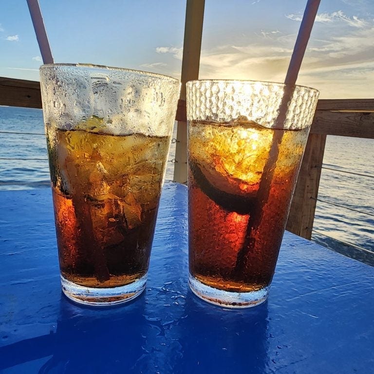 Two glasses of beer with condensation are placed on a wooden surface outdoors at sunset, overlooking the ocean in Roatan, Honduras.