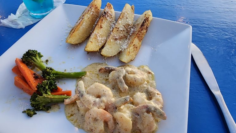 A plate of fried potato wedges, steamed vegetables, and creamy shrimp pasta is served on a white plate at a dining area in Roatan, Honduras, with a blue table and a glass of blue-colored beverage nearby.