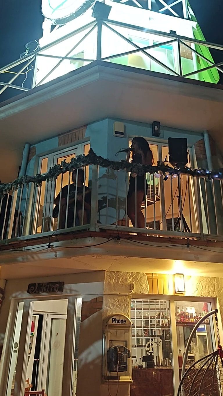 A woman performs karaoke on a balcony of a building in Roatan, Honduras, with a music stand and microphone, illuminated by warm interior lighting at night.