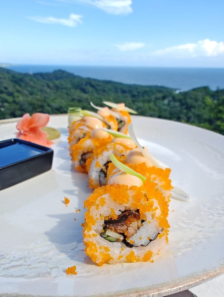 Sushi rolls topped with orange fish roe and drizzled with sauce are presented on a white plate with a scenic view of lush green hills, the ocean, and a blue sky in Roatan, Honduras.