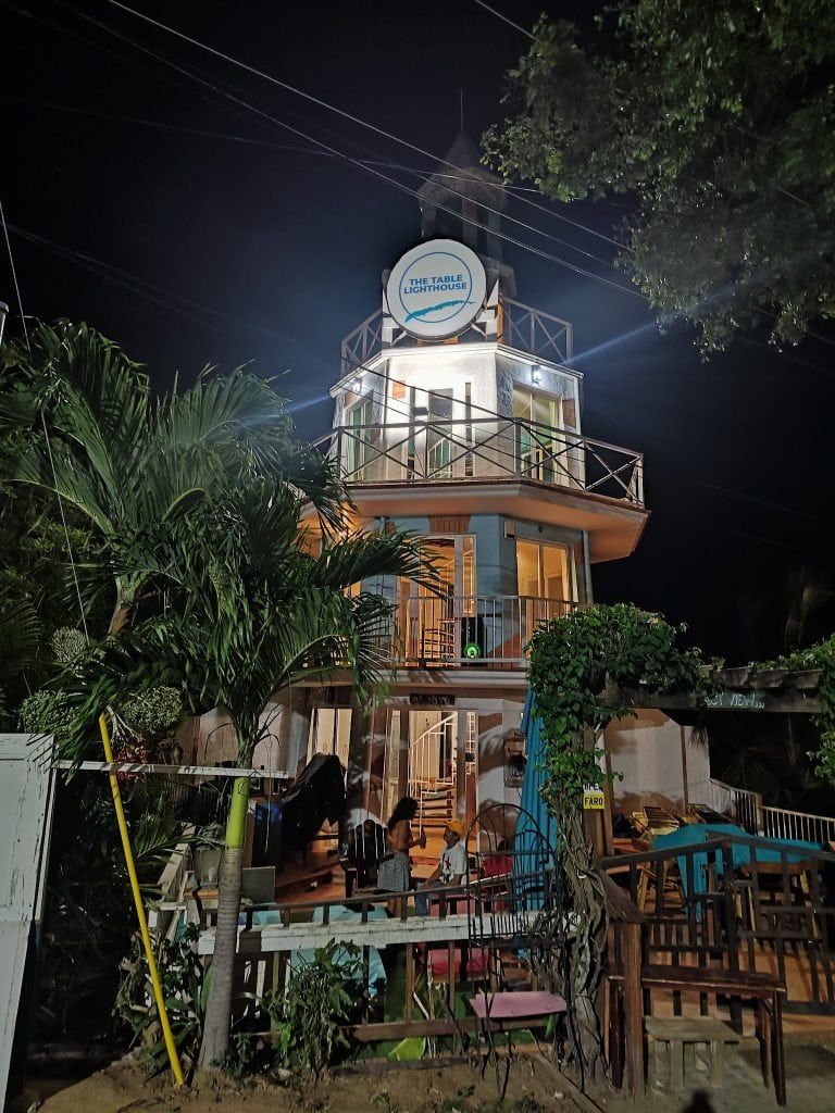 The image shows a multi-story restaurant or business building at night in Roatan, Honduras, with a sign that reads "The Table Lighthouse," surrounded by lush green trees and illuminated windows, with outdoor seating and people gathered in front.