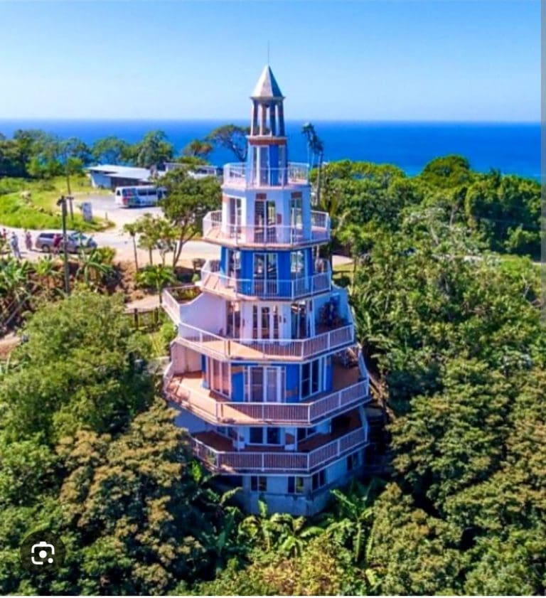 A tall, multi-level lighthouse with multiple decks and glass windows sits among lush green trees on Roatan, Honduras, with the ocean visible in the background under a clear blue sky.