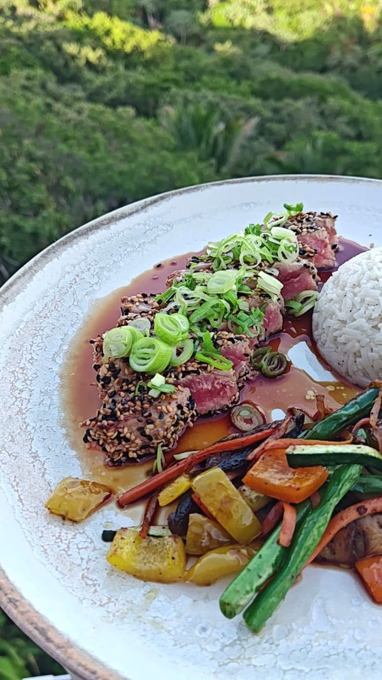 A plate of seared tuna coated with sesame seeds, topped with sliced green onions, served with roasted vegetables and white rice, with lush greenery in the background on Roatan, Honduras.