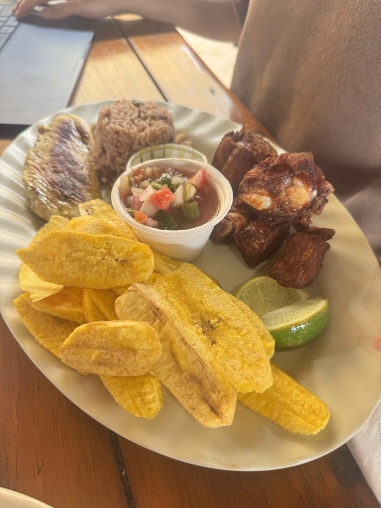 A plate of traditional Honduran cuisine served in Roatan, featuring fried plantains, rice and beans, crispy pork ribs, a bowl of vegetable salsa, and lime wedges, set on a wooden table in Honduras.