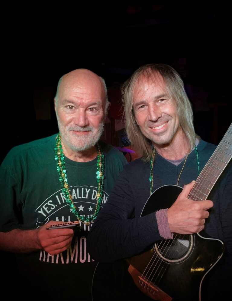 Two men with beards and long hair are smiling while playing acoustic guitars at a lively indoor event in Roatan, Honduras, wearing necklaces and casual clothing.