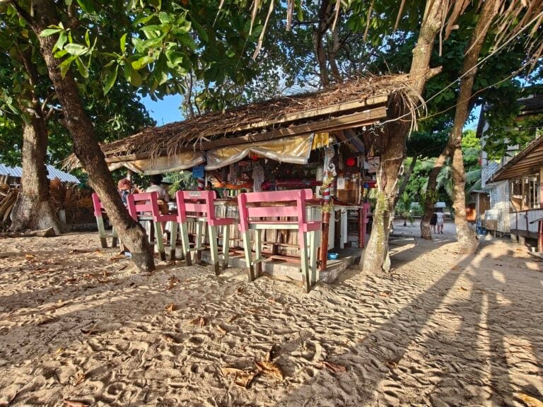 An outdoor beachside bar in Roatan, Honduras, featuring a thatched roof, pink bar stools, and surrounded by trees with sandy ground and a clear blue sky.
