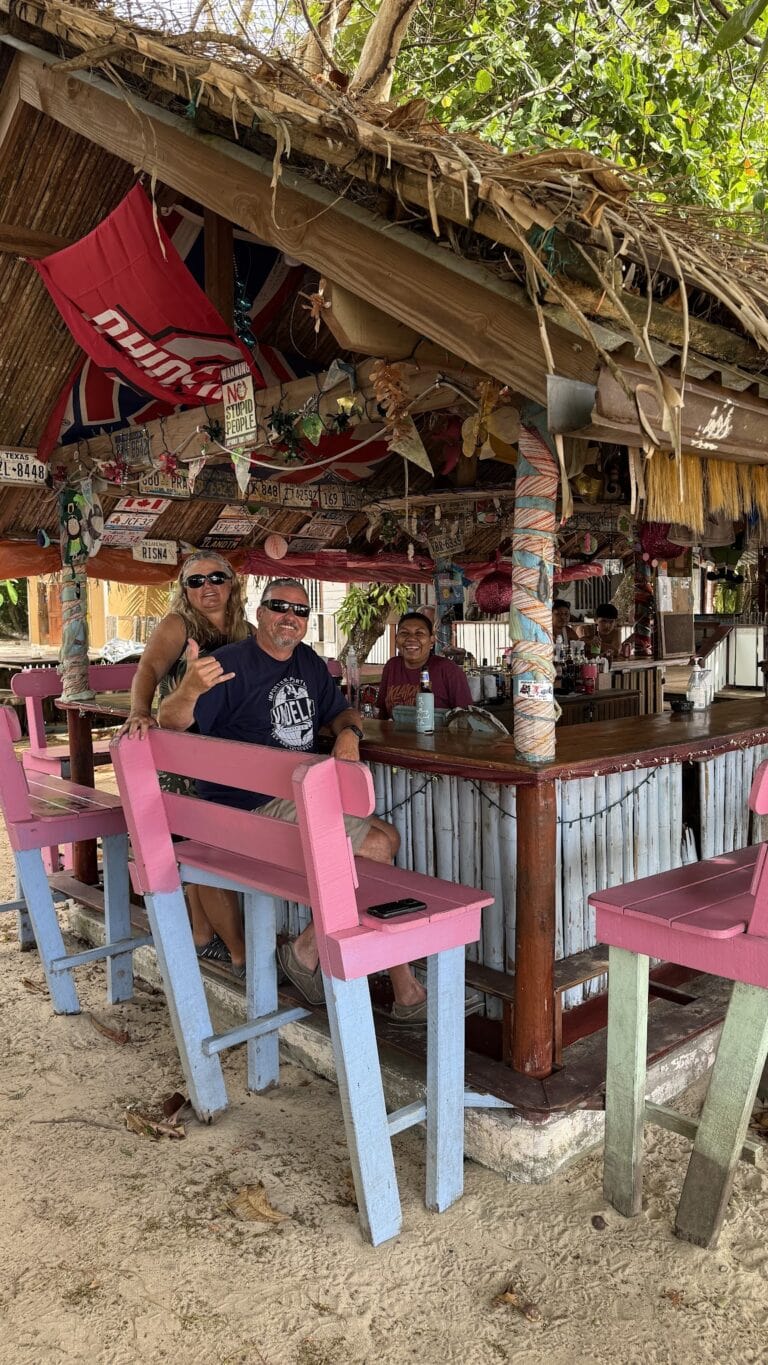 A group of three people enjoying drinks at a colorful, open-air bar with a thatched roof decorated with license plates and tropical ornaments on Roatan, Honduras.