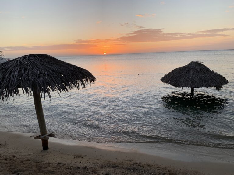 A sunset over the calm waters of Roatan, Honduras, with two thatched umbrellas in the foreground on a sandy beach.