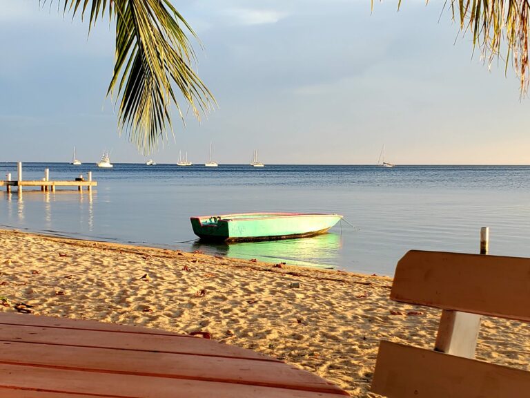 A small green and white boat rests on the sandy beach near calm ocean waters in Roatan, Honduras, with sailboats anchored in the distance under a partly cloudy sky.