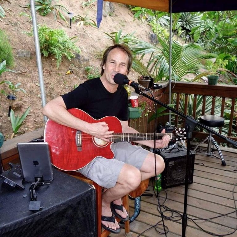 A man playing an acoustic guitar and singing into a microphone on a wooden outdoor deck surrounded by lush greenery in Roatan, Honduras.