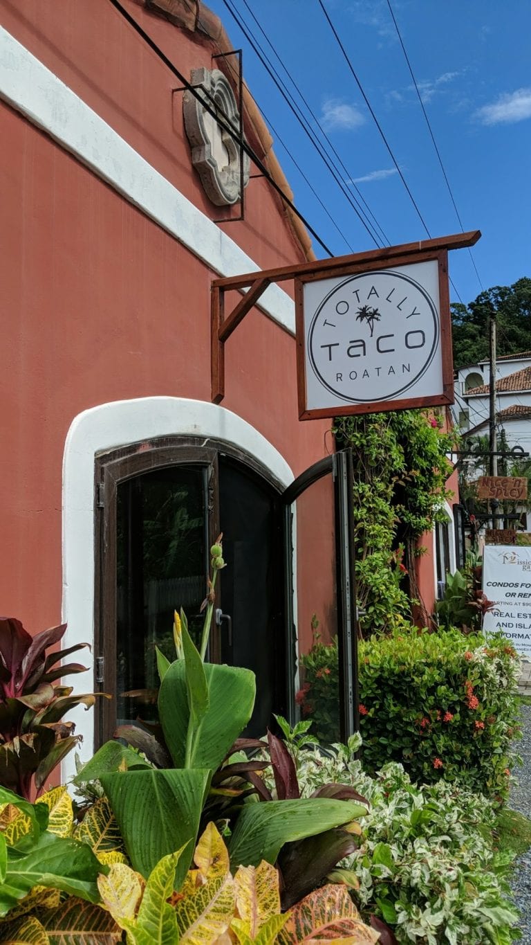 A storefront in Roatan, Honduras, features a white sign with black lettering reading "Totally Taco Roatan" and a palm tree logo, set against a coral-colored building with surrounded by lush green and flowering plants.