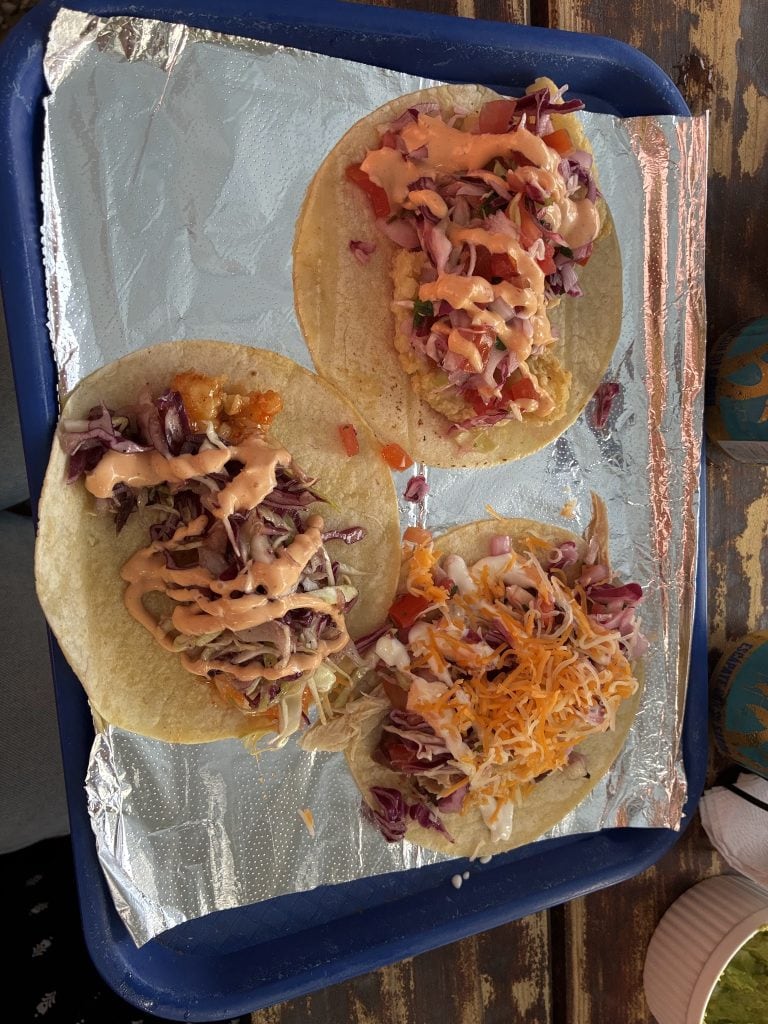 Three tacos filled with shredded cabbage, diced tomatoes, and creamy sauce are served on a foil-lined tray at a food stand in Roatan, Honduras.