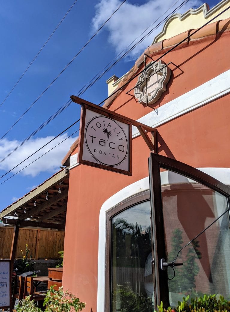 A peach-colored building with a large window, a decorative crest above the entrance, and a sign reading "Totally Tacos Roatan" hangs outside, with an outdoor seating area, plants, and a wooden fence under a blue sky in Roatan, Honduras.