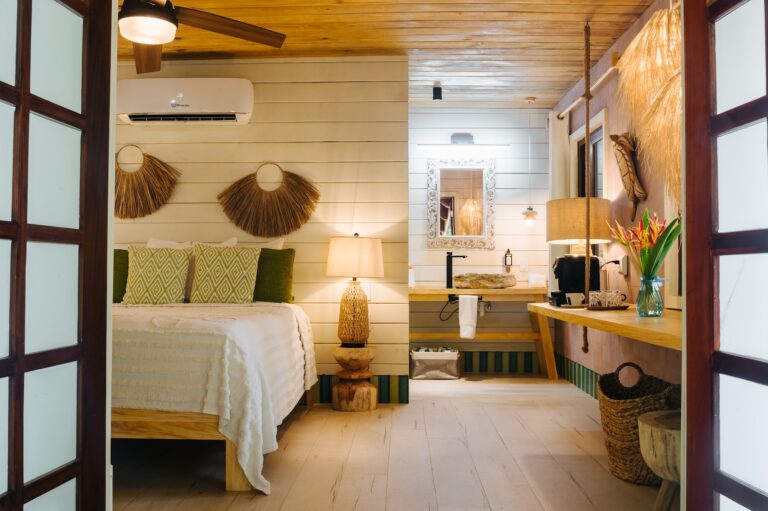 A cozy bedroom in a Roatan, Honduras vacation rental features a bed with green patterned pillows, a wooden nightstand with a wicker lamp, and decor including straw wall hangings, with a view into a small bathroom area that has a rustic wooden sink and a mirror.